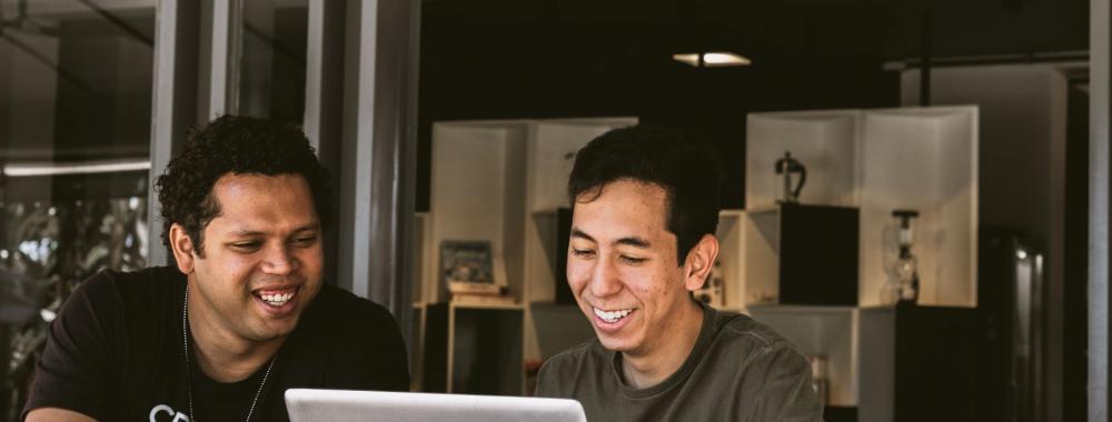 Two men smile while working together on a laptop