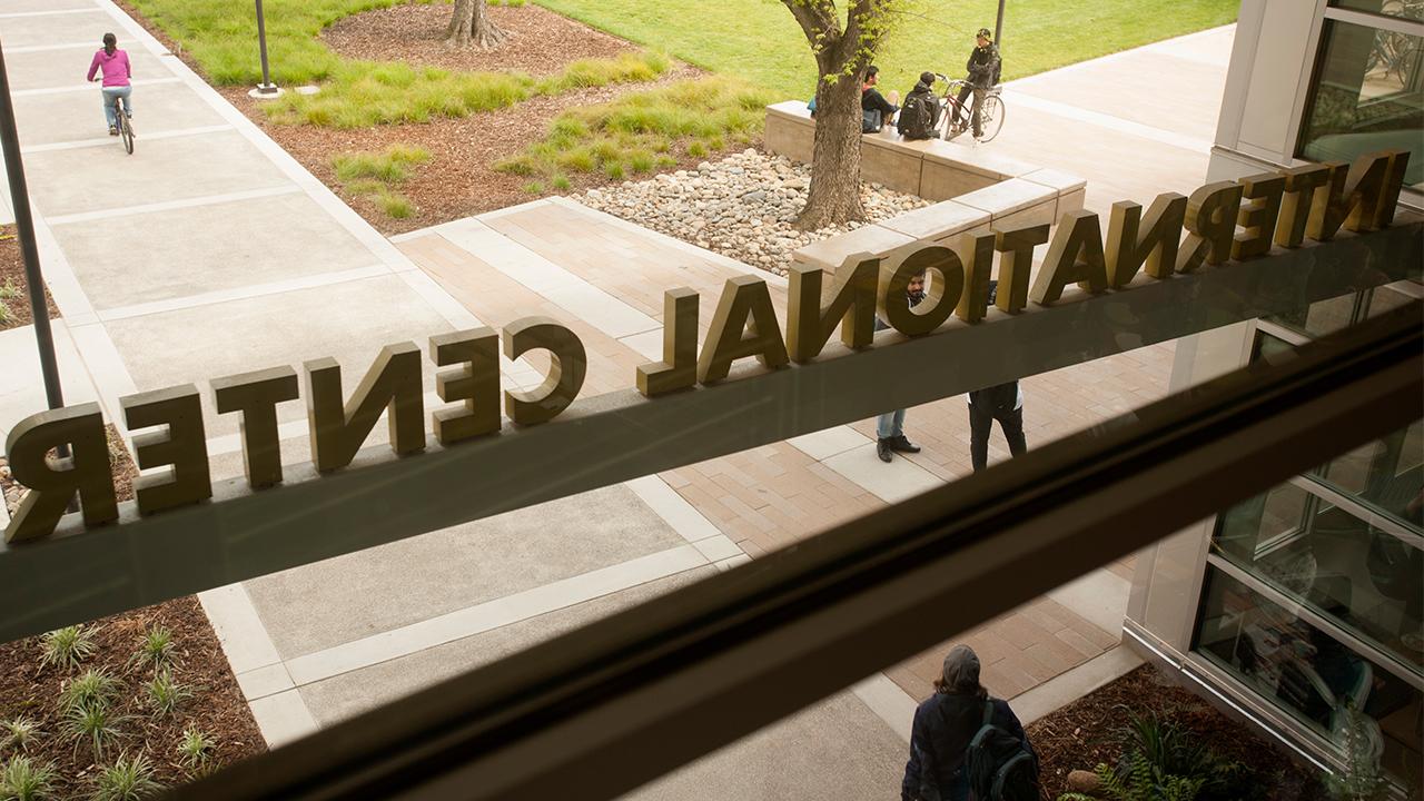 A photo from inside the International Center on the UC Davis campus looking out the main entrance window, taken on a fall day. The International Center sign is backwards from this vantage point looking out as a student wearing a hat, blue jacket and backpack walks away from the doors. Three other students, one sitting on a bike and two others sitting, father under a tree outside. A student in a pink sweater rides her bike away from the building.