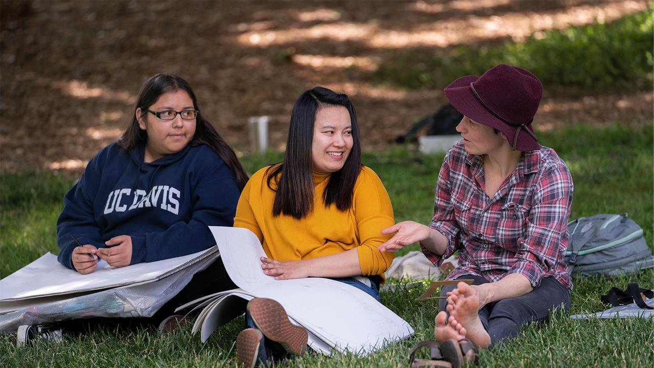 Three young women sit on the grass at the Arboretum on the UC Davis campus. The woman on the left has long straight dark hair and wears glasses and a navy UC Davis hoodie. She has a large sketch pad on her lap. The woman in the middle has long straight black hair worn in a half ponytail. She wears a yellow sweater and has a large sketch pad on her lap. The woman on the right has a maroon wool hat, a deep pink flannel shirt and sits cross legged with her shoes off. She talks to the other two women.