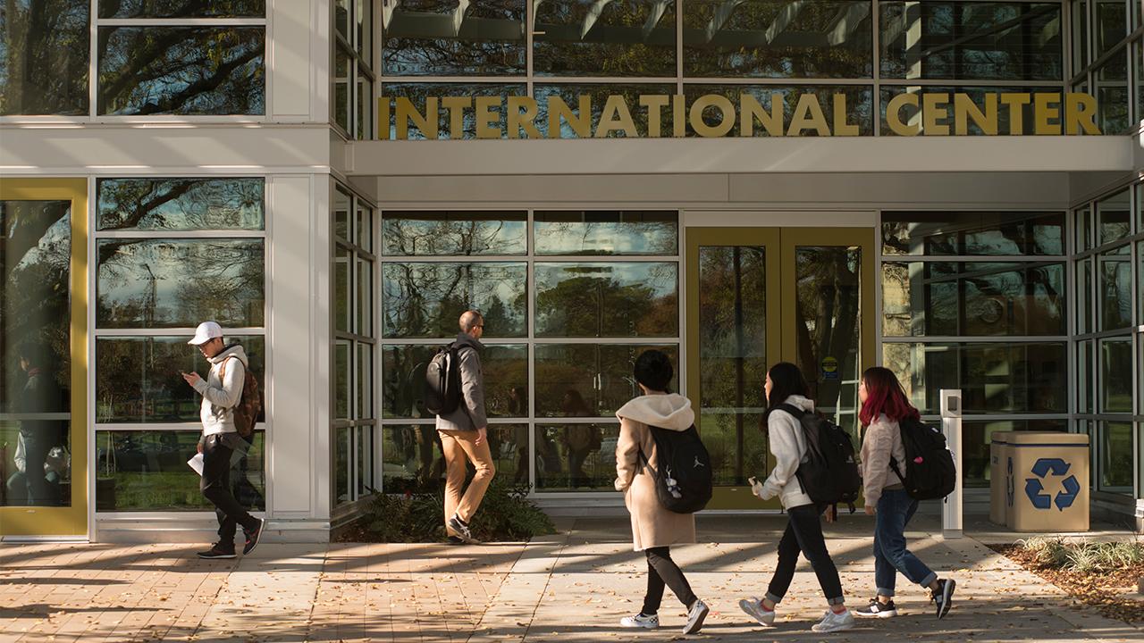 On a sunny day, three female students dressed for cool weather and wearing backpacks approach the doors of the International Center on the right side of the photo. One male student wearing a jacket and backpack approaches from the left, and one male students looks at his phone as he walks away from the building at the very left of the photo.