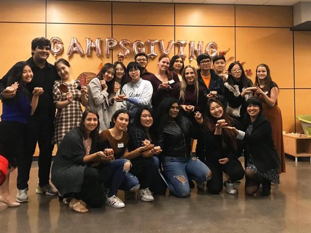 A group of 20 mentors from the Global Ambassador Mentorship Program pose inside the lobby of the UC Davis International Center. Fourteen students (four men and ten women) stand in a row at the back, and six women kneel in the front row. They hold small pumpkins and are dressed for fall weather.