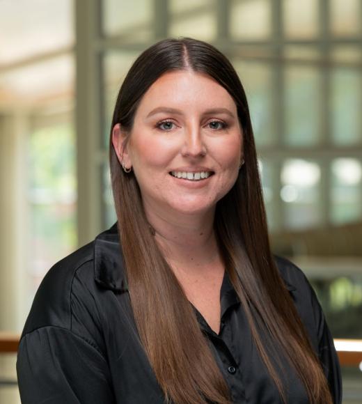 Marissa Hoggard stands in the International Center at UC Davis. She wears a black blouse. Her brown hair is long and worn down, and she smiles at the camera. 