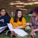 Three young women sit on the grass at the Arboretum on the UC Davis campus. The woman on the left has long straight dark hair and wears glasses and a navy UC Davis hoodie. She has a large sketch pad on her lap. The woman in the middle has long straight black hair worn in a half ponytail. She wears a yellow sweater and has a large sketch pad on her lap. The woman on the right has a maroon wool hat, a deep pink flannel shirt and sits cross legged with her shoes off. She talks to the other two women.