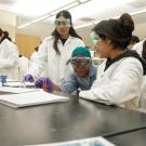 In a chemistry lab, a TA with teal hair leans in to examine the measurement of a test tube as the two students around her watch and interact with her. They all wear lab coats and protective eye goggles. 