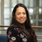 Lorena Galvan poses smiling at the camera for a headshot inside the International Center with windows in the background. She has long dark hair and wears a black top with large flowers. 