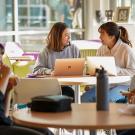 Inside the lobby area of the International Center with daylight coming in through the glass windows. Two students out of focus in the foreground sit opposite from each other at a table and are talking. One is eating. In the background in focus, two other students sit next to each other, both working on silver laptops. They smile at each other.
