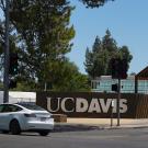The UC Davis sign on a concrete wall on the corner of College Park and Howard Way at one of the entrances to the UC Davis campus on a sunny day. Three students on foot cross the street in the crosswalk toward the sign and another student walks a bike across the crosswalk away from the sign. A white Tesla crosses the street heading east past the sign.
