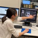 Karen wears a face covering and lab coat. She is seated at a desk with computer screens and she points to a small graph on one screen while two colleagues look over her shoulder at the screen.