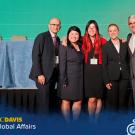 Bob Kiaii stands in front of a stage with three woman and one man all wearing business attire. The stage behind them is set up for a panel of speakers with draped tables and chairs in front of a large light green screen. A graphic with the UC Davis Global Affairs logo and #GlobalAggies lays over the photo at the bottom.