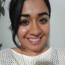 Banicia Chatkara smiles in a selfie. She sits against a white wall indoors. Her black hair is pulled back and side swept bangs hang down to the length of her chin on her right. She wears a white sweater, a diamond or crystal stud in her nose on her left, and a delicate silver chain necklace.