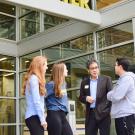 Four people stand outside the main entrance of the International Center at UC Davis in the shade. The first woman has long strawberry blonde hair and wears black pants and a blue collared shirt. A second woman has long light brown hair and wears a UC Davis Global Affairs dark heather blue T-shirt and black jeans. Next to her, Wesley Young wears a gray blazer over a light blue collared shirt and navy pants. He wears dark rimmed glasses and looks at the young man to his left who wears a light gray hoodie.