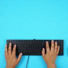hands on a black computer keyboard with a black mouse to the right. The keyboard and mouse sit on a bright blue table top.