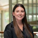 Marissa Hoggard stands in the International Center at UC Davis. She wears a black blouse. Her brown hair is long and worn down, and she smiles at the camera. 