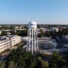 Aerial view of UC Davis campus looking north from the south side of campus. The main focus is on a white water tower with blue writing that reads "UC DAVIS". Mrak Hall rises in the distance to the right of the photo and it is a clear day with a bright blue sky.