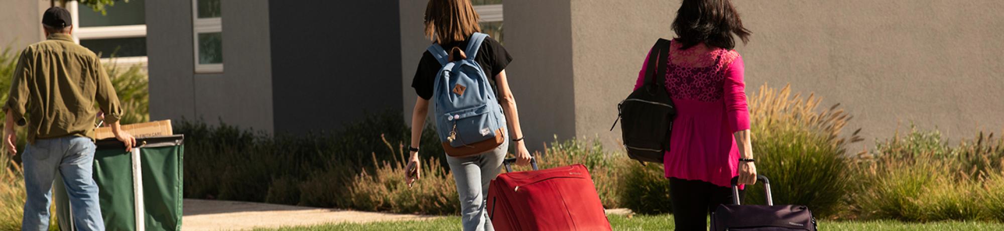 Students carry bags and roll red and black suitcases along the walkway during student move-in on the UC Davis campus. A man with a large cart walks in the distance ahead of them.