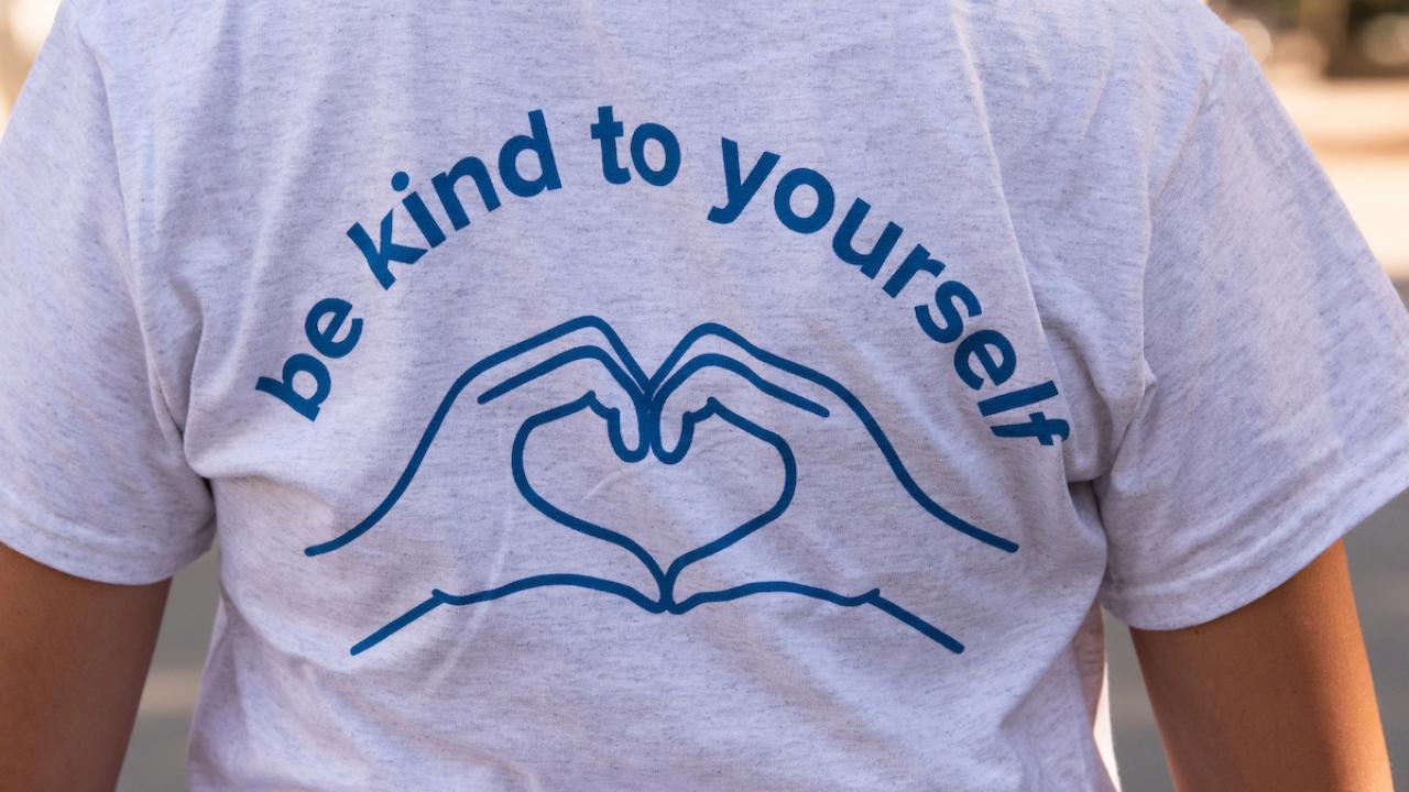 The back of a student wearing a light gray T-shirt with a blue linear outline of two hands cupped to form a heart and above the words "Be Kind to Yourself."