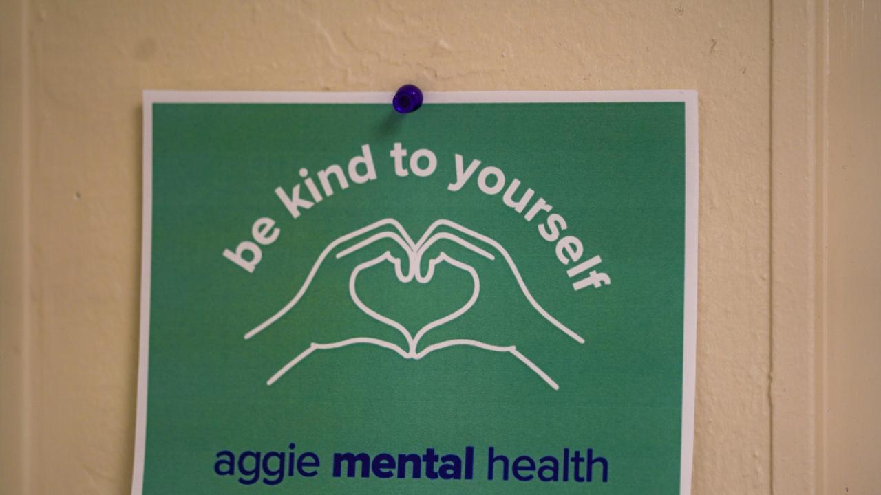 A sign on a wall says "be kind to yourself" and "aggie mental health." I also has a line illustration of two hands making a heart shape