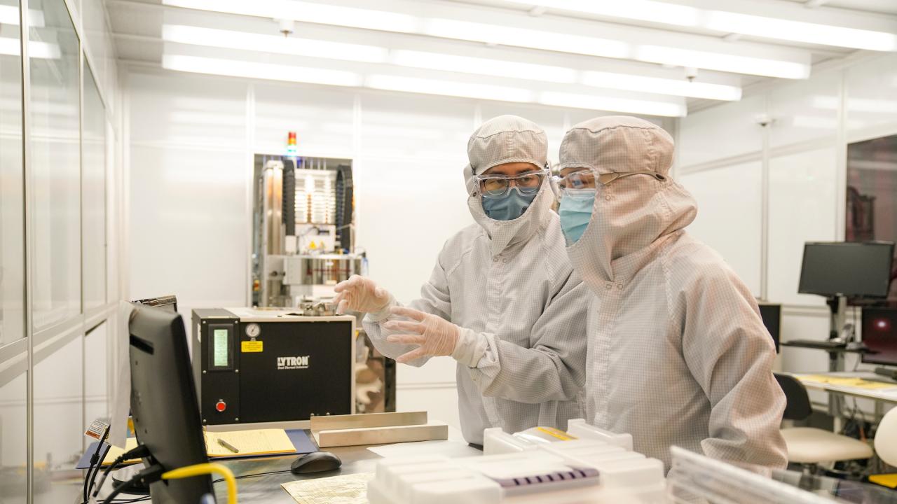 Two technicians in full cleanroom suits and masks pointing at a computer in a sterile lab.