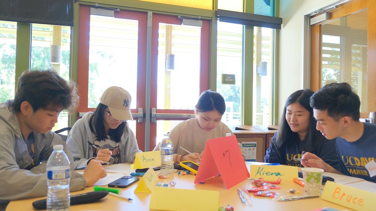 Five students sit at a table with notes and materials spread out in front of them with their heads down in collaboration.