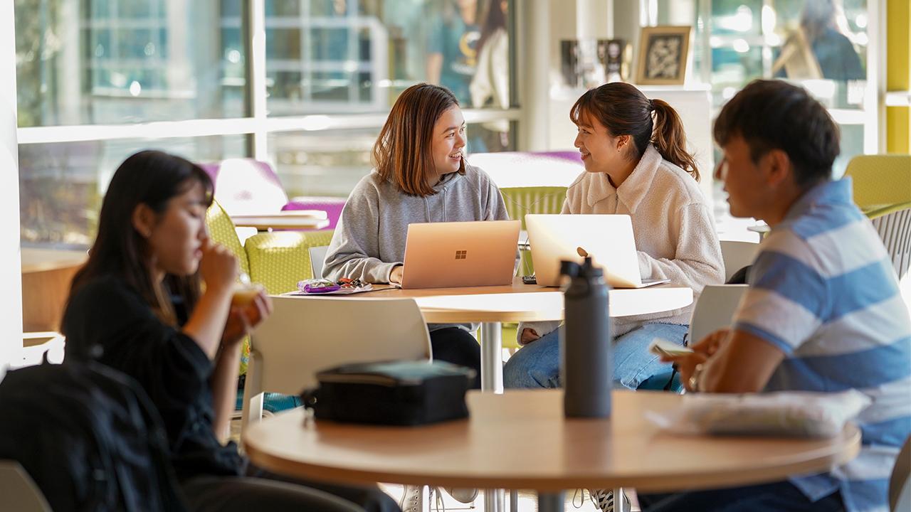 Inside the lobby area of the International Center with daylight coming in through the glass windows. Two students out of focus in the foreground sit opposite from each other at a table and are talking. One is eating. In the background in focus, two other students sit next to each other, both working on silver laptops. They smile at each other.