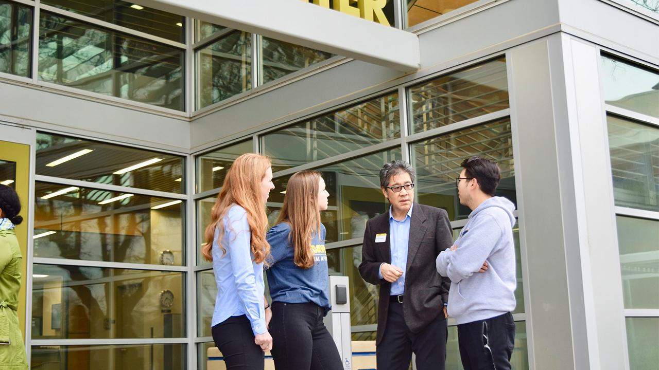 Four people stand outside the main entrance of the International Center at UC Davis in the shade. The first woman has long strawberry blonde hair and wears black pants and a blue collared shirt. A second woman has long light brown hair and wears a UC Davis Global Affairs dark heather blue T-shirt and black jeans. Next to her, Wesley Young wears a gray blazer over a light blue collared shirt and navy pants. He wears dark rimmed glasses and looks at the young man to his left who wears a light gray hoodie.