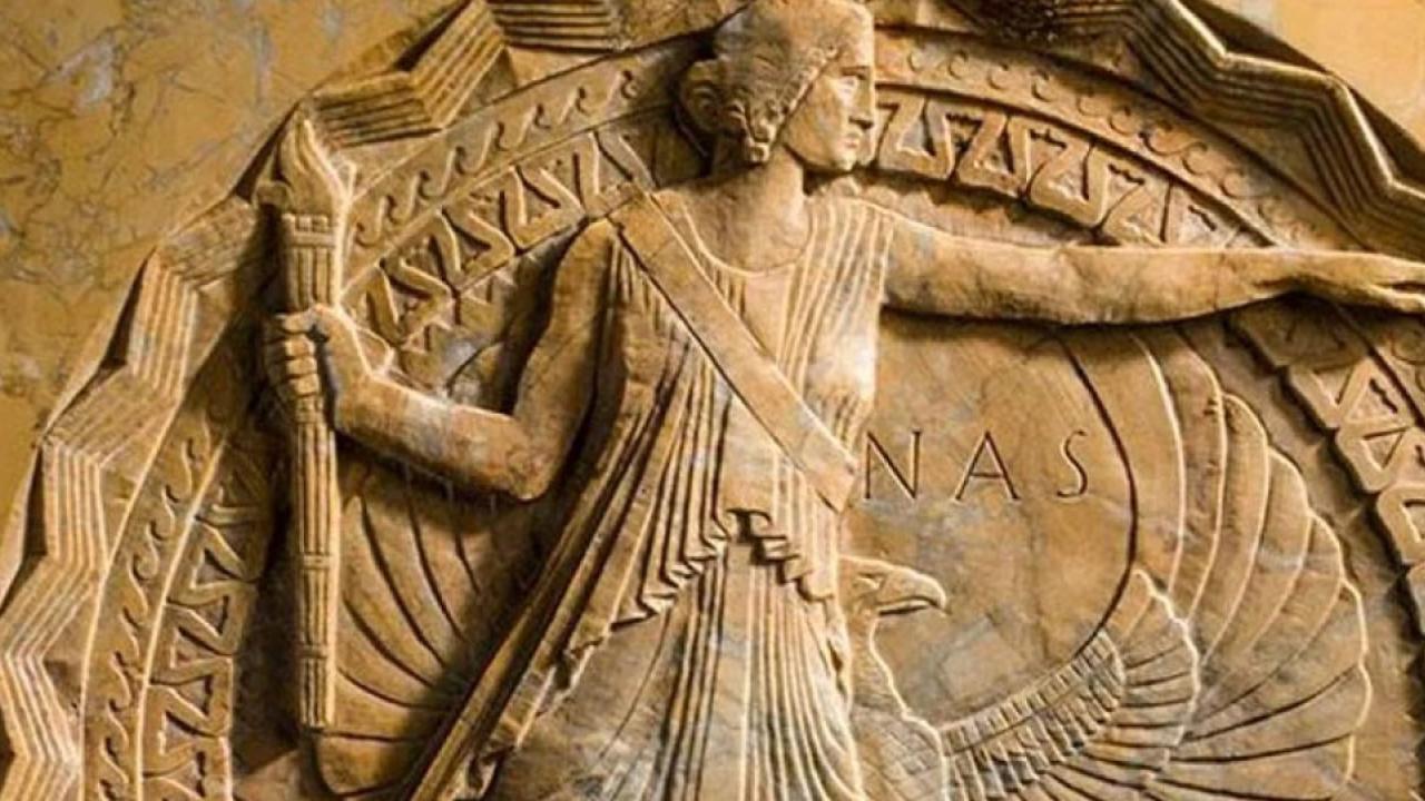 The National Academy of Sciences logo made from a light stone features a figure wearing a robe, their arm extended to the right and on the left side, holding a torch.