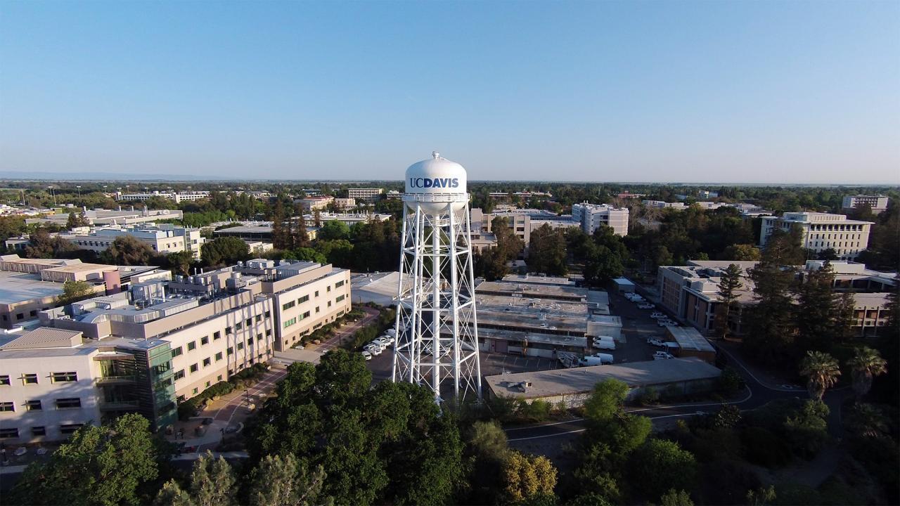 Aerial view of UC Davis campus looking north from the south side of campus. The main focus is on a white water tower with blue writing that reads "UC DAVIS". Mrak Hall rises in the distance to the right of the photo and it is a clear day with a bright blue sky.