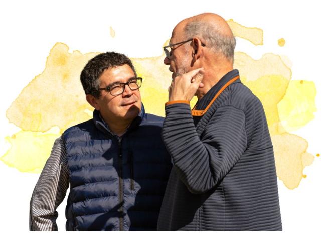 Two men engaging in conversation, one wearing a vest, against a colorful background.