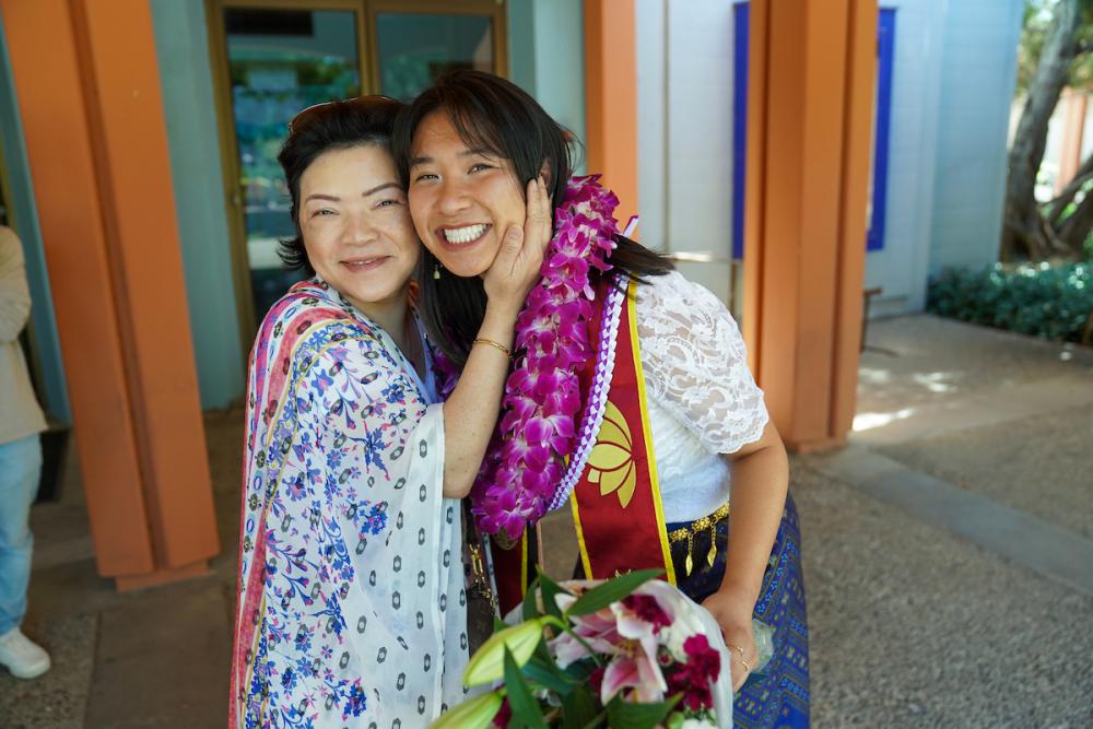 A mother embraces her child, who wears their graduation stole, a lei and holds a bouquet of flowers. Both smile at the camera.