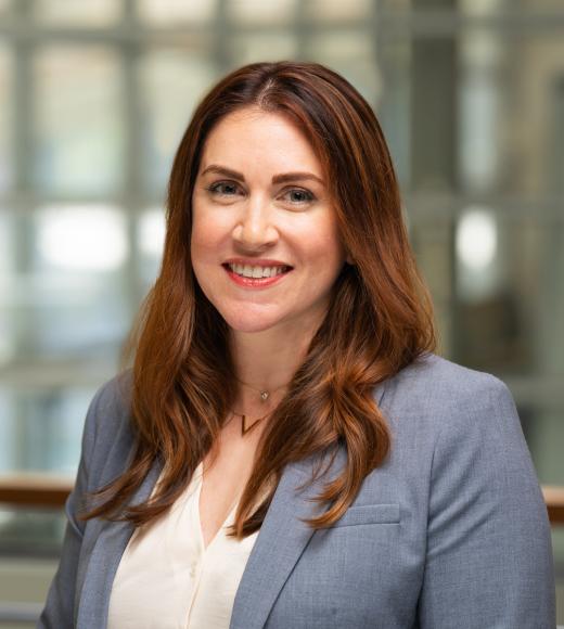 Jana smiles in a headshot with the windows of the International Center in the background. She wears a gold necklace, gray/blue blazer and cream top. Her auburn hair is worn down over her shoulders.