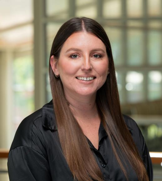 Marissa Hoggard stands in the International Center at UC Davis. She wears a black blouse. Her brown hair is long and worn down, and she smiles at the camera.