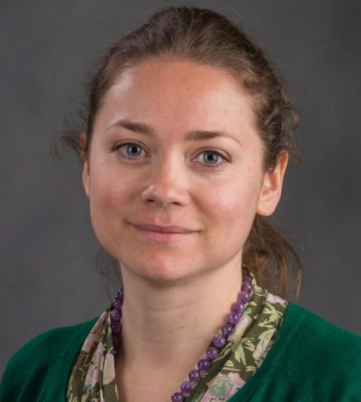 Headshot of Lena Zadorozhna in front of a gray backdrop. She wears an emerald green sweater over a green and pink floral blouse with a amethyst beaded necklace. Her light brown hair is pulled back in a low ponytail.