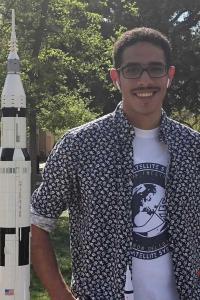 Abdallah Hashem stands in front of a replica of a NASA rocket. He wears a black button-up collared shirt with a light white floral pattern worn with rolled cuffs and open and a T-shirt with the seal of a satellite system on the front in black. He smiles at the camera. His black hair is textured and worn short, and he has AirPods in his ears and wears black rectangular-framed glasses.