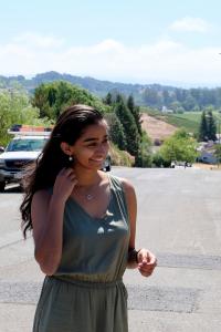Aditi Mahajan photographed outdoors on a street smiling and looking off to the right. She wears an olive green sleeveless dress, a silver pendant necklace and silver earrings. Her long black hair is worn down, and there is a parked truck and tree-covered green hills in the background behind her.