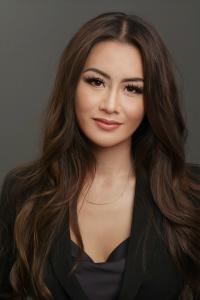 Arista Wu smiles slightly at the camera. She stands in front of a dark gray background. Her long dark hair is worn straight and falls over her shoulders. She wears a black blouse under a black blazer, and a delicate gold necklace. 