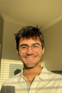 Cem Nesiri stands in the sunlight indoors. He smiles at the camera. His dark hair is short and he wears round wireframed glasses, and a light blue collared shirt.