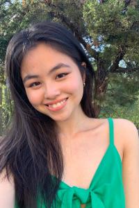 Eindra Khine smiles in a selfie taken in the natural light outdoors against trees as the backdrop. She wears a green sleeveless blouse with a bow on the front, small gold hoop earrings, and her black hair is worn straight and long over her right shoulder.