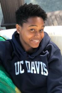 Haley Ali wears a navy UC Davis hoodie with the words "UC Davis" in white lettering across the chest. She lounges in a white chair and smiles at the camera. Her head is turned slightly to her left, revealing a silver stud earring in her right ear. Her short black hair is longer on top and closely cropped on the sides.