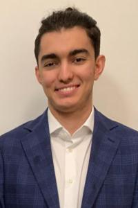 Hashem Bkayrat stands against a white background as he smiles at the camera. He wears a blue checked blazer over a white collared shirt. His dark hair is groomed short.