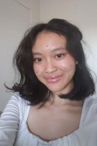 Helena Ngo smiles in a selfie taken indoors in the natural light with a white wall behind her. Her dark hair is worn straight and reaches her collarbone. She wears a white scoop-neck top. 