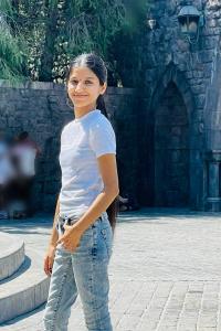 Mangurleen Kaur smiles slightly to the camera and stands in profile in front of a stone castle wall. She wears a white T-shirt and acid wash blue jeans. Her black hair is long and straight, and hangs down her back in a low ponytail.