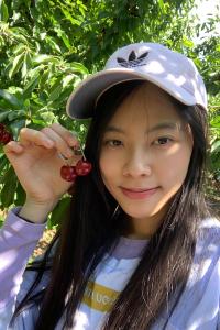 Minnie Yu stands in the shade under a tree holding up two cherries connected at the stem. She wears a white Adidas baseball cap with a black logo, and a lavender jacket over a white T-shirt. She smiles slightly and has long straight black hair that falls over her shoulders.