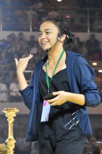 Margaret Castañeda Lee photographed smiling on stage at an event. She wears her black hair pulled back in a low bun, a blue jacket over a black jumpsuit, a green lanyard, and a microphone headset over her left ear.
