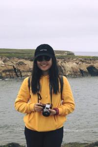 Odelia Chong stands at cliffs overlooking the ocean on an overcast day. She holds a camera that hangs around her neck, and wears round sunglasses, a black baseball cap that says in white lettering "I can't even," and a yellow hoodie.