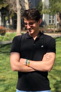 Pavel Martinez stands with his arms casually crossed, looking down to his right and smiling. He is outside in the sun with grass, trees, and a white building behind him in the background. He wears sunglasses, a black polo shirt, and two woven bracelets on his left wrist, one green and the other yellow and green. His brown hair is worn neatly styled and long on the top. 