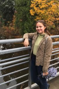 Rachelle Taveau smiles as she stands on a bridge overlooking a creek with fall foliage changing behind her along the water. Her right arm rests on the top of the metal bridge railing, nearly the height of her shoulder. She wears a light green turtleneck under a khaki colored jacket, and dark blue jeans, and she holds a closed royal blue and white hounds tooth umbrella. She has shoulder length light brown hair that is pulled back into a half ponytail 