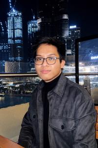 Ritz Tolentino sits overlooking a night city skyline with skyscrapers in the background. He wears his black hair cropped close at the sides and longer on top. He wears a black shirt under a black leather jacket. 