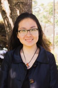Vy Nguyen stands under a tree during the daytime. She smiles at the camera. Vy has long straight black hair, and she wears rectangular glasses, a multi-colored pendant on a black silk cord around her neck, a black top, and a rich eggplant purple jacket over it with large square bronze buttons. 
