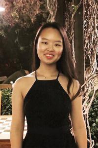 Youngyun Kim smiles as she stands outdoors at night. She wears a sleeveless black dress and a delicate gold necklace. Her long black hair is work straight and down.