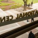 A photo from inside the International Center on the UC Davis campus looking out the main entrance window, taken on a fall day. The International Center sign is backwards from this vantage point looking out as a student wearing a hat, blue jacket and backpack walks away from the doors. Three other students, one sitting on a bike and two others sitting, father under a tree outside. A student in a pink sweater rides her bike away from the building.